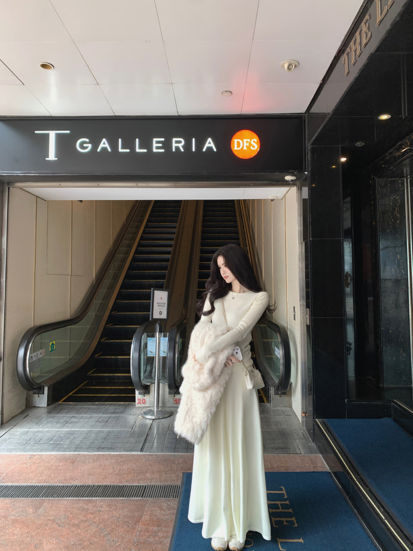 Woman wearing a long cream dress and holding a white fur coat, standing in front of a T GALLERIA DFS store entrance with escalators.