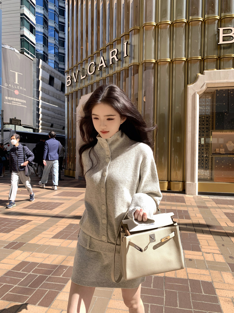 Woman wearing a beige knit cardigan and matching skirt, holding a cream handbag, standing in front of a BVLGARI store.
