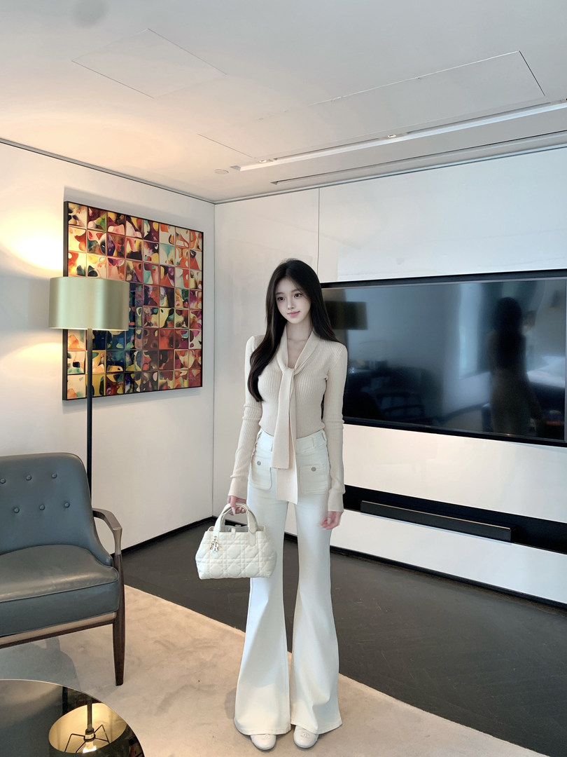 Woman wearing cream ribbed top with tie neck, white flared pants, and holding white quilted handbag in modern living room