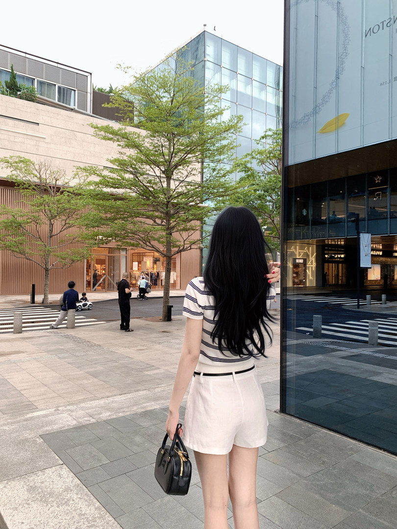 Woman with long black hair wearing striped top and white shorts, holding black handbag, walking outdoors in urban setting