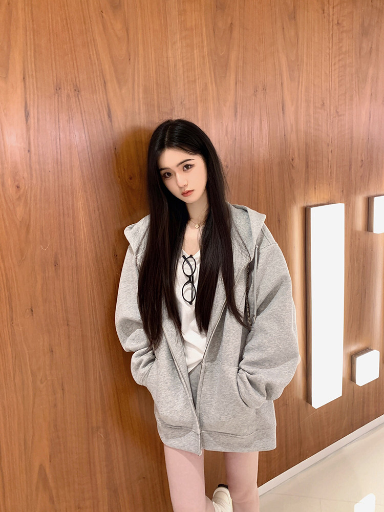 Woman wearing a gray zip-up hoodie over a white top, standing against a wooden wall