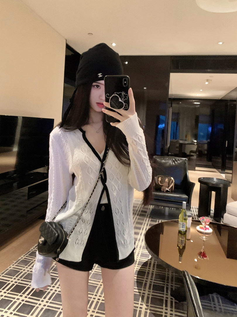 Woman taking mirror selfie wearing white knit cardigan, black shorts, and black beanie with Chanel logo
