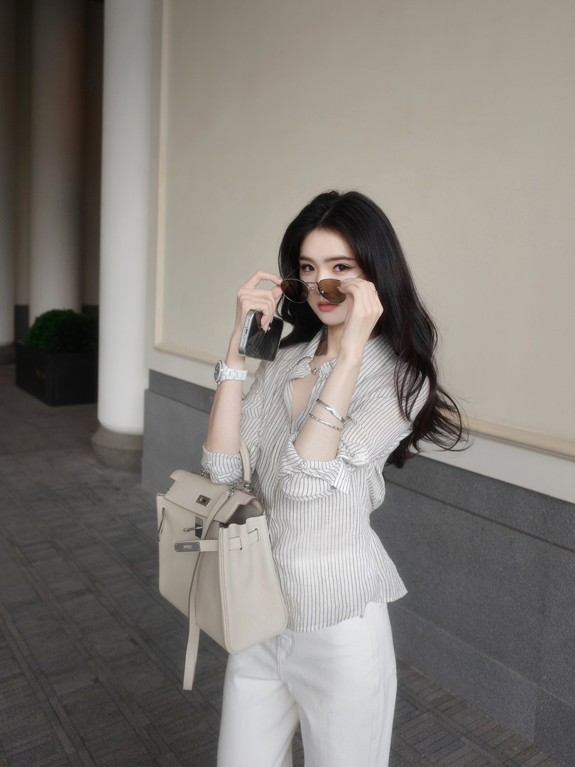 Woman wearing striped blouse and white pants, holding sunglasses and a beige handbag
