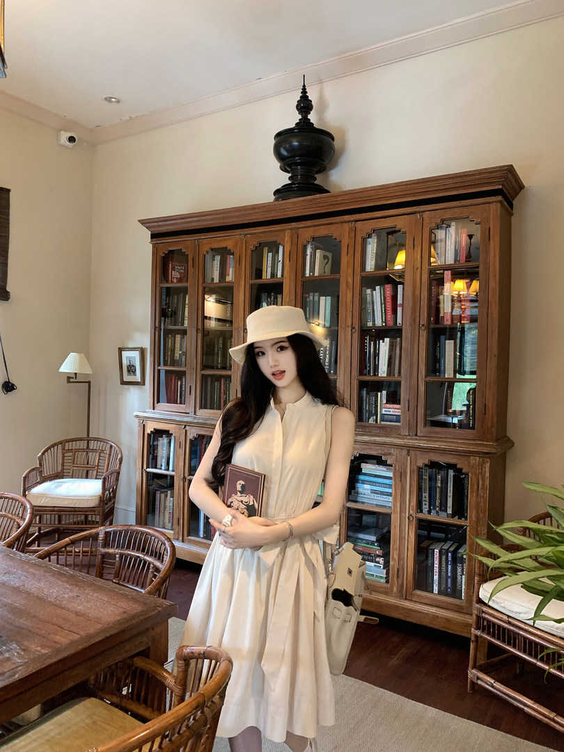 Woman wearing a white sleeveless dress and hat, standing in a room with bookshelves.