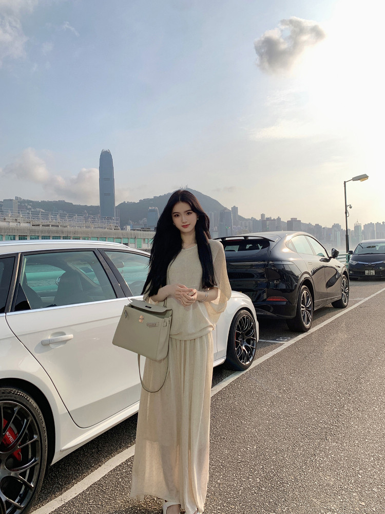 Woman wearing beige long dress and holding handbag, standing in parking lot with city skyline background
