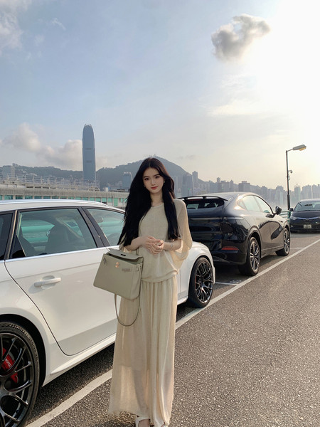 Woman wearing beige long dress and holding handbag, standing in parking lot with city skyline background