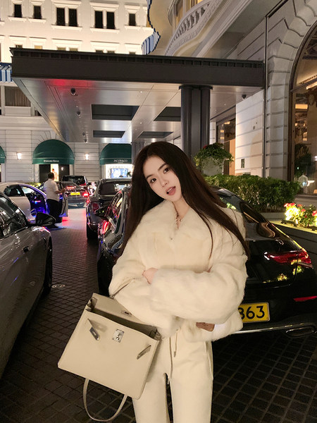 Woman wearing a white fluffy coat and holding a beige handbag in front of a luxury hotel entrance.