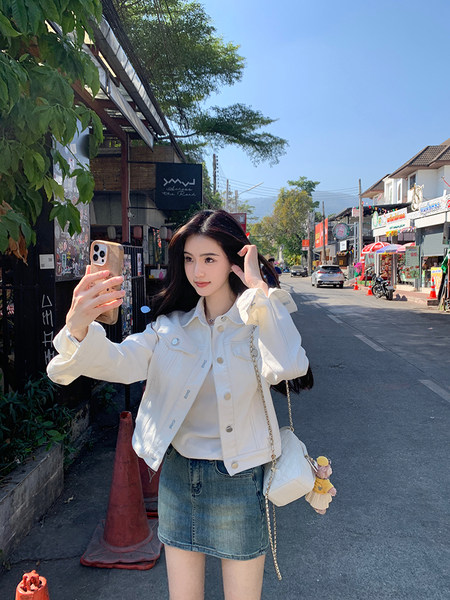 Woman wearing white denim jacket and denim skirt taking a selfie on a street
