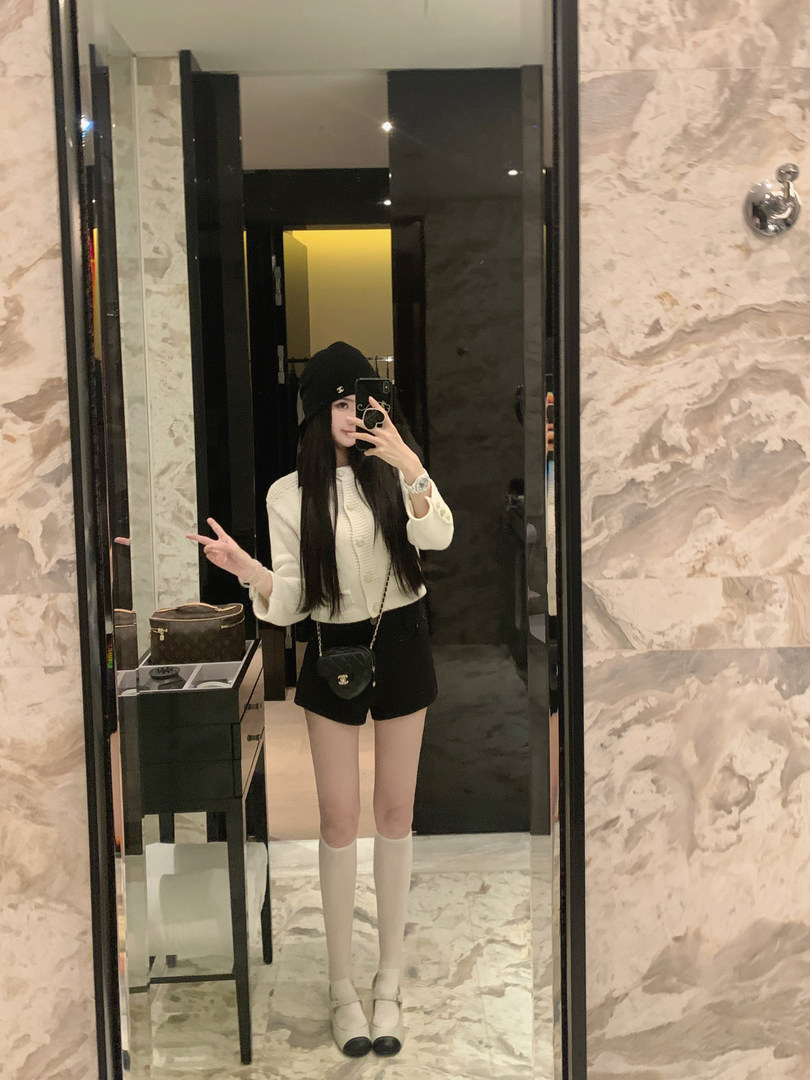 Mirror selfie of a woman wearing a white cropped cardigan, black shorts, beanie, knee socks, and Mary Jane shoes