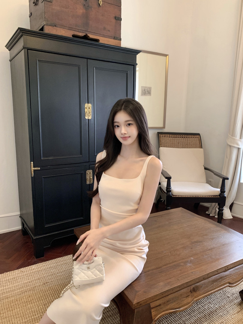 Woman wearing a beige sleeveless dress while sitting on a wooden table with a small white handbag.