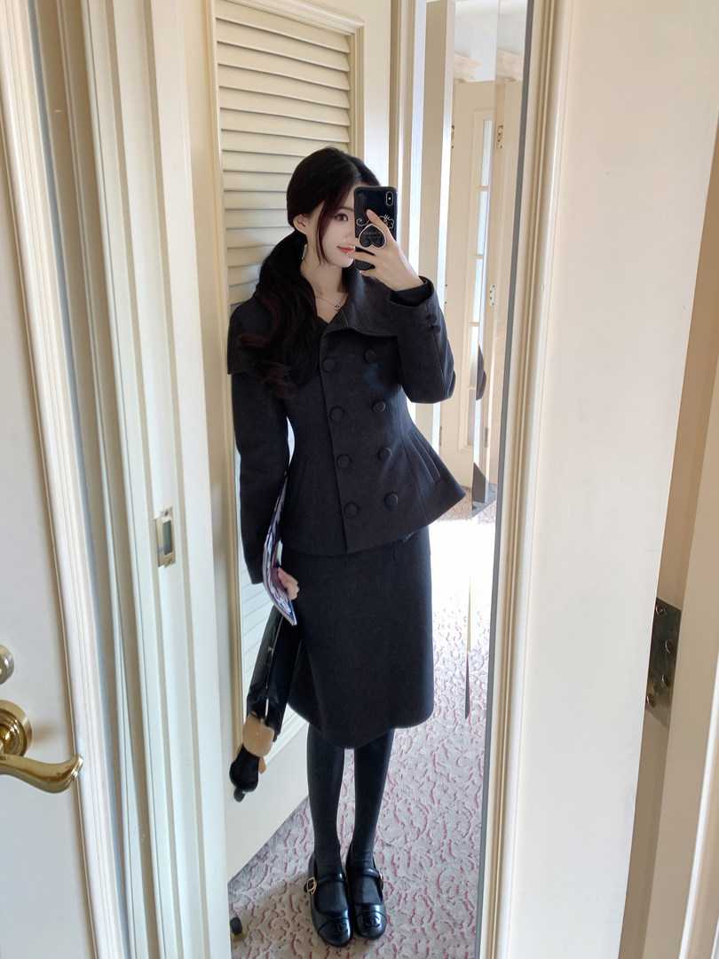 Woman taking mirror selfie wearing a black double-breasted peplum coat with matching skirt, black tights, and loafers