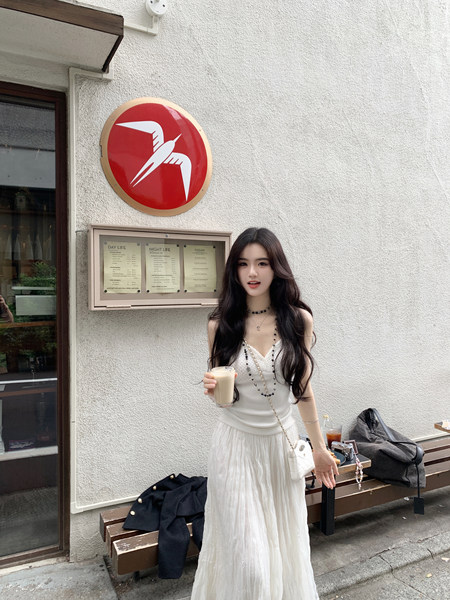 Woman wearing a white ribbed knit top and pleated skirt, holding a drink, standing outdoors near a cafe menu board.