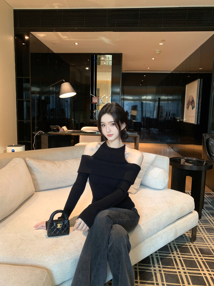Woman wearing a black off-shoulder ribbed top and gray pants, sitting on a beige couch with a small black handbag.
