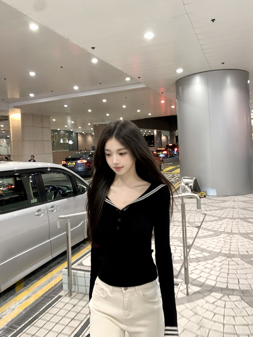 Woman wearing black long-sleeve top with white trim and white pants, standing outdoors near cars