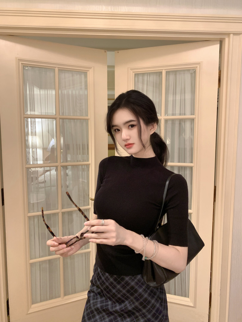 Woman wearing a black ribbed top and plaid skirt, holding glasses, with a black bag over her shoulder.