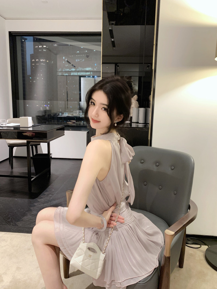 Woman wearing a light purple sleeveless dress with bow detail, sitting on a gray chair with a white quilted handbag.