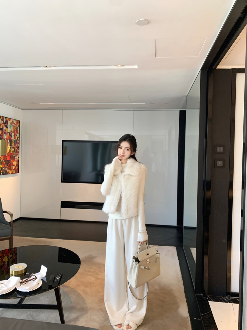 Woman wearing a white fur-trimmed coat and wide-leg pants, holding a handbag in a modern living room