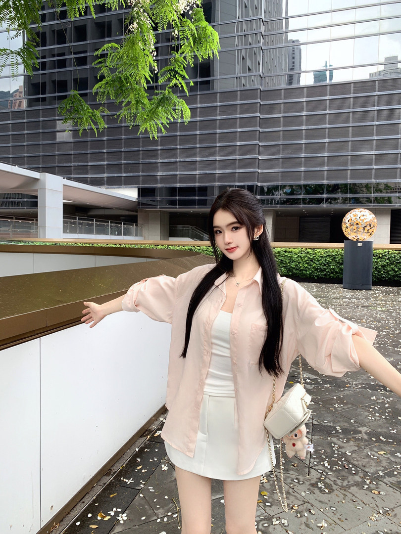 Woman wearing a light pink oversized shirt over a white top and white shorts, standing outdoors with a modern building background.