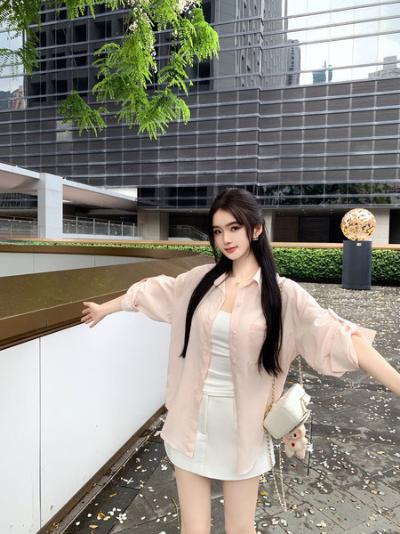 Woman wearing a light pink oversized shirt over a white top and white shorts, standing outdoors with a modern building background.