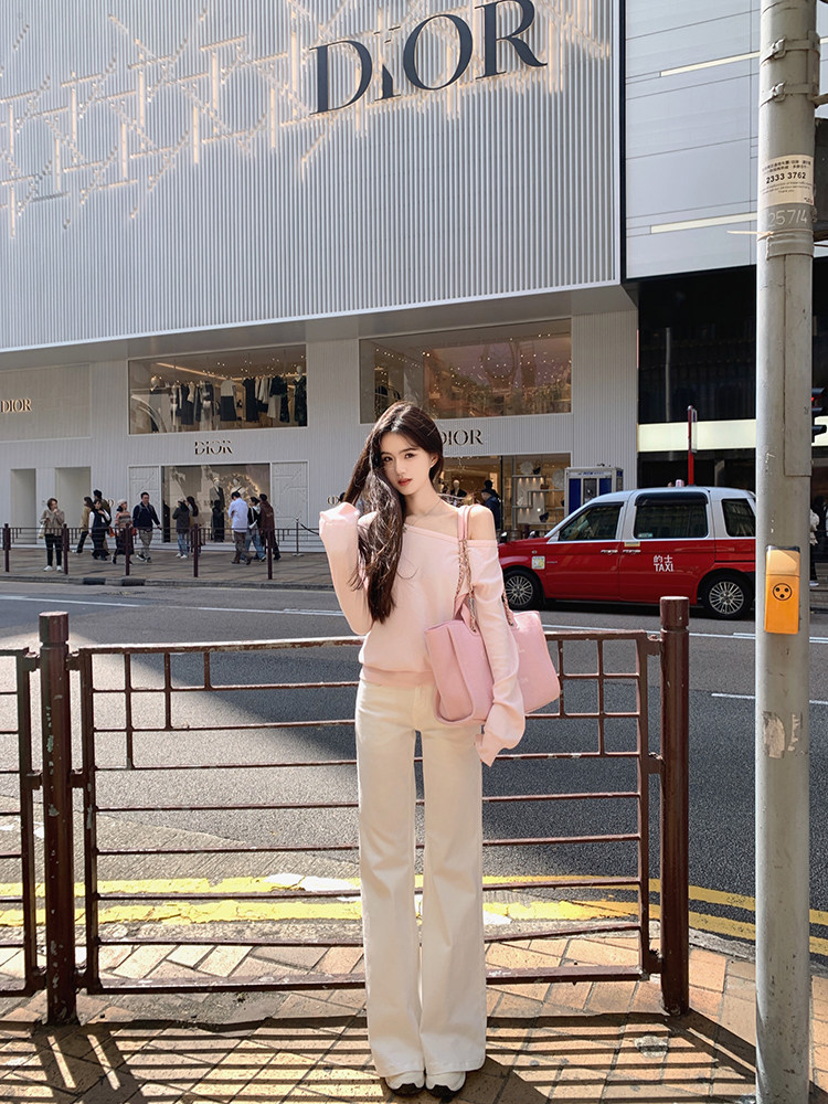 Woman wearing pink off-shoulder top and white pants with a pink bag, standing on a street in front of a Dior store.