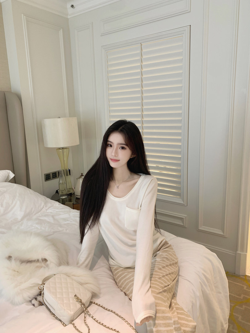 Woman wearing a white long-sleeve top and striped pants sitting on a bed with a white quilted handbag.
