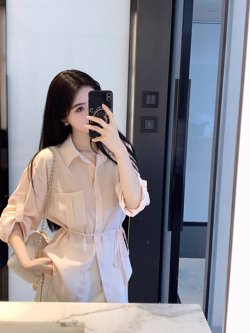 Woman taking mirror selfie wearing a light pink button-up shirt dress with rolled sleeves and a belt.