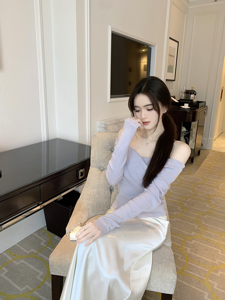 Woman wearing a light purple off-shoulder top and white satin skirt, seated on a beige armchair in a luxury room.