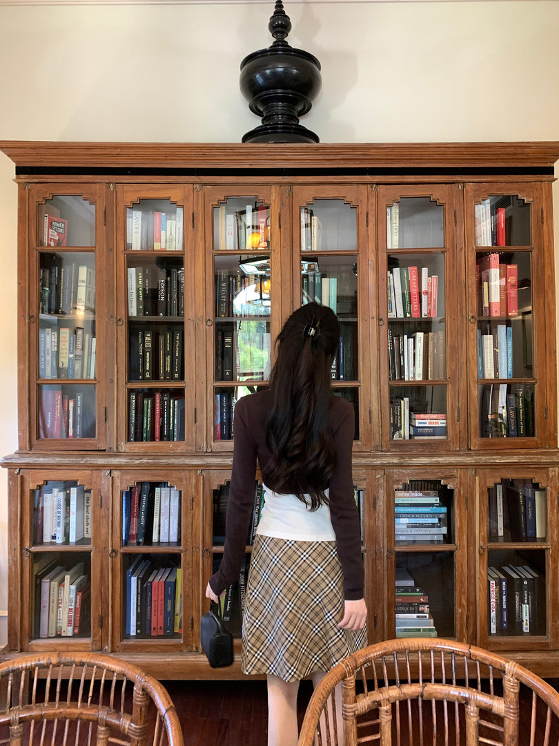 A woman wearing a plaid skirt and brown top, standing in front of a wooden bookshelf.