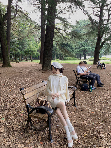 Woman wearing a cream knit sweater dress, white hat, and white shoes sitting on a park bench
