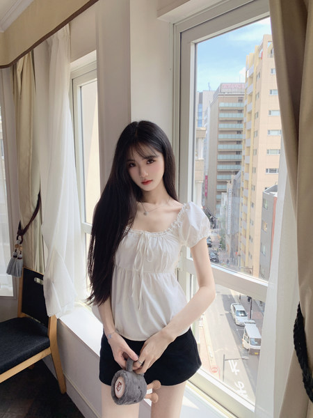Woman wearing white puff-sleeve top and black shorts by a window with city view