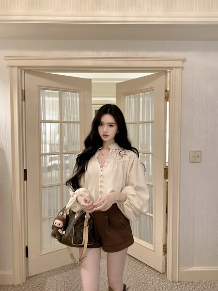 Woman wearing a cream blouse with lace collar and brown shorts, holding a patterned handbag with a plush toy attached