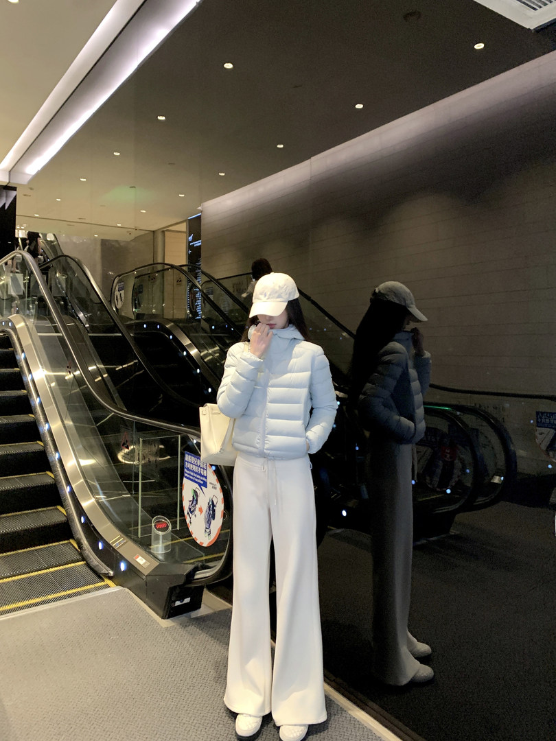 Woman wearing light blue puffer jacket and white wide-leg pants in a mall near escalators