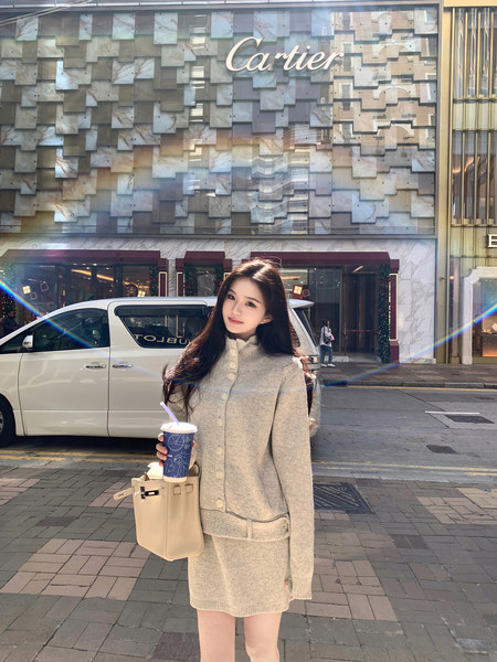 Woman wearing a beige knit cardigan and matching skirt set, holding a drink and tote bag, standing in front of a Cartier store.