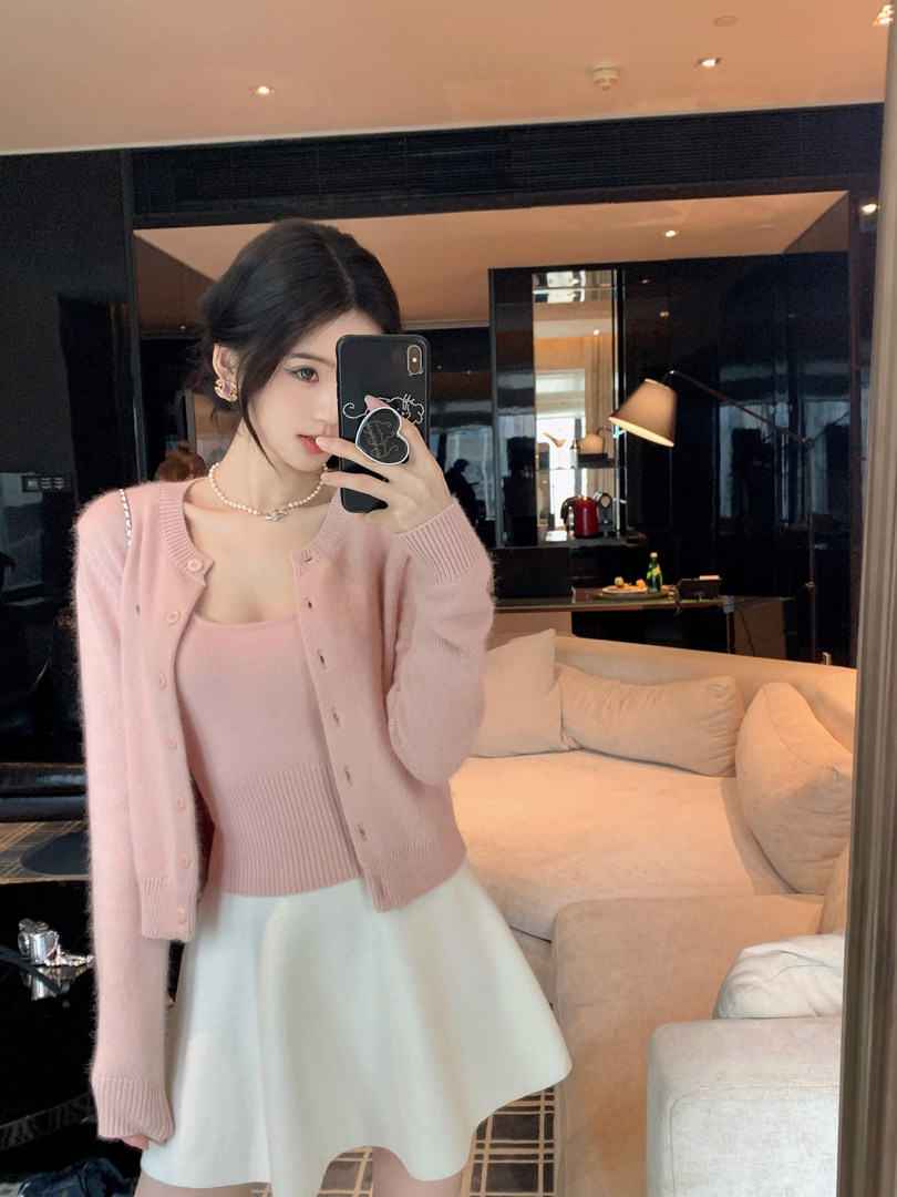Woman wearing a pink knit cardigan and white skirt, taking a mirror selfie