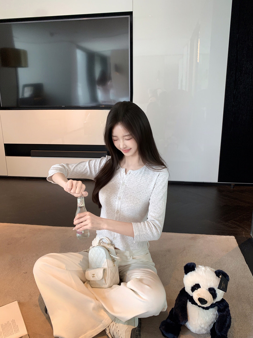 Woman sitting on floor wearing white patterned long-sleeve top and wide-leg pants, holding a bottle, with a small quilted handbag and stuffed panda beside her