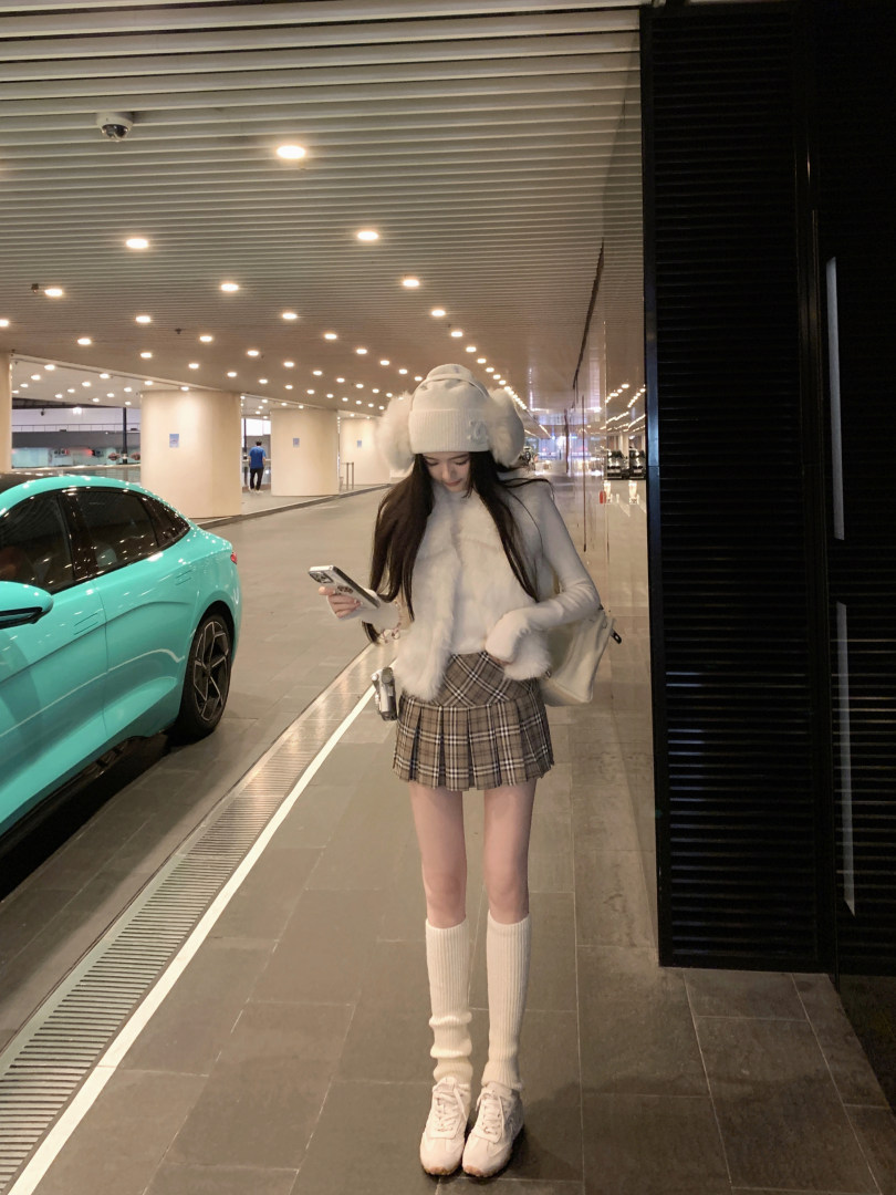 Woman wearing white knit hat with fluffy ears, white fuzzy vest, plaid pleated skirt, white knee-high socks, and white sneakers, standing in a parking garage