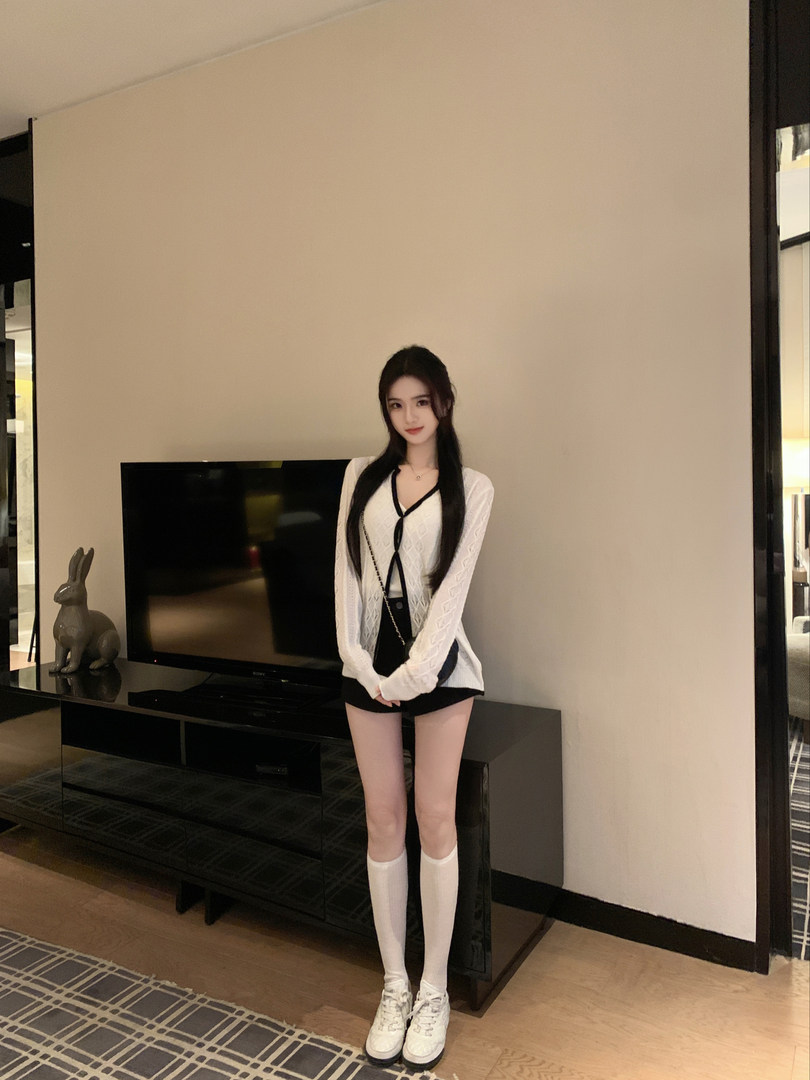 Woman wearing a white knitted cardigan, black shorts, and white knee-high socks with sneakers.