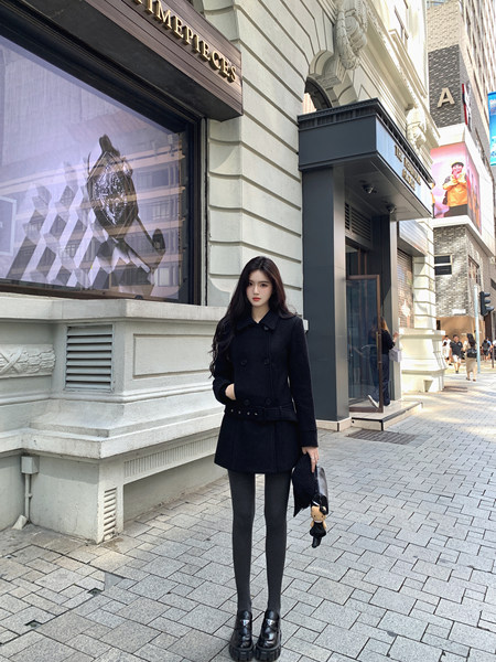 Woman wearing black coat, black tights, and black platform shoes standing on a city street