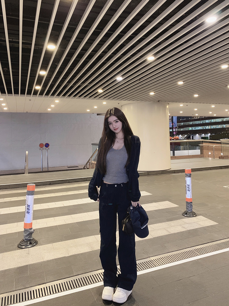 Woman wearing a gray top, dark jacket, and wide-leg pants, holding a bag, standing in an urban setting.