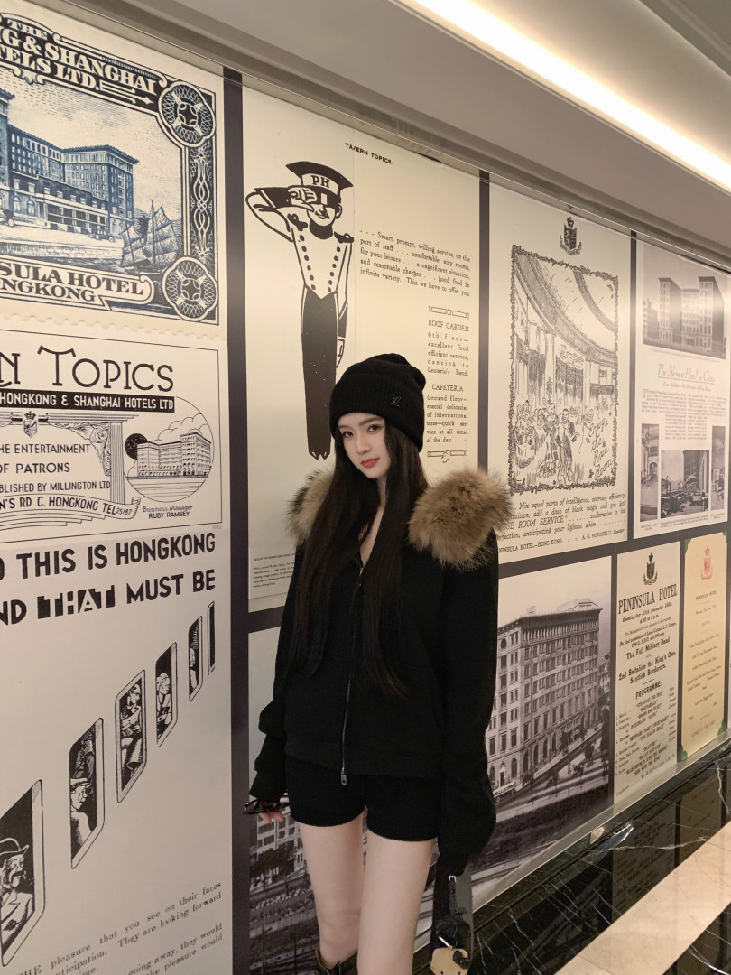 Woman wearing black jacket with fur hood and black beanie standing in front of vintage hotel posters