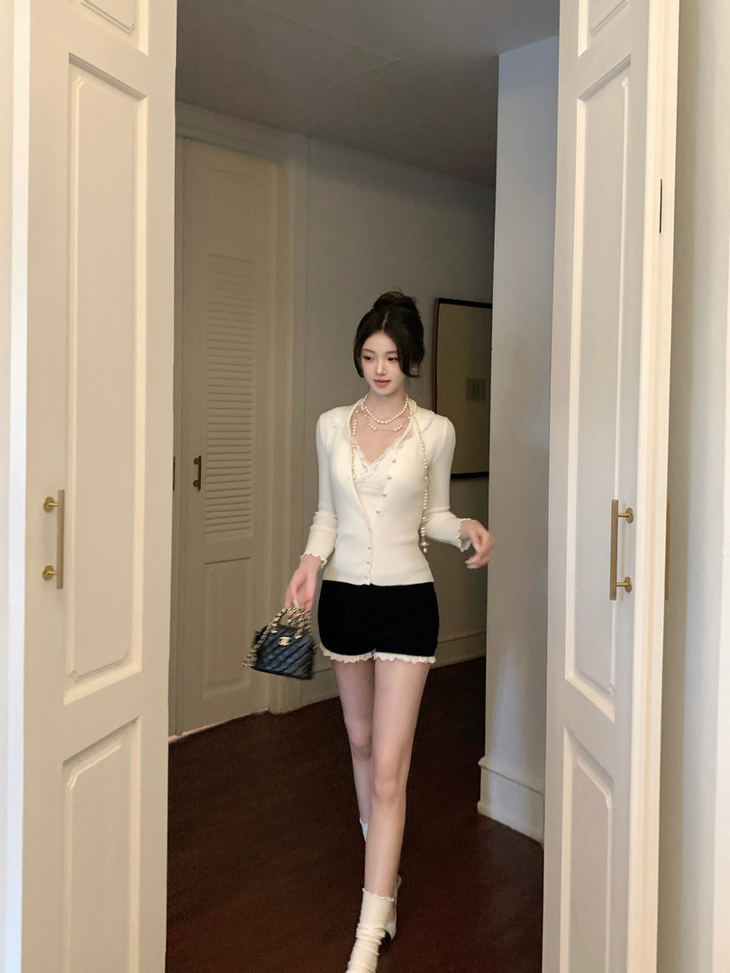 Woman wearing a white cardigan with pearl details, black shorts, and holding a small handbag.