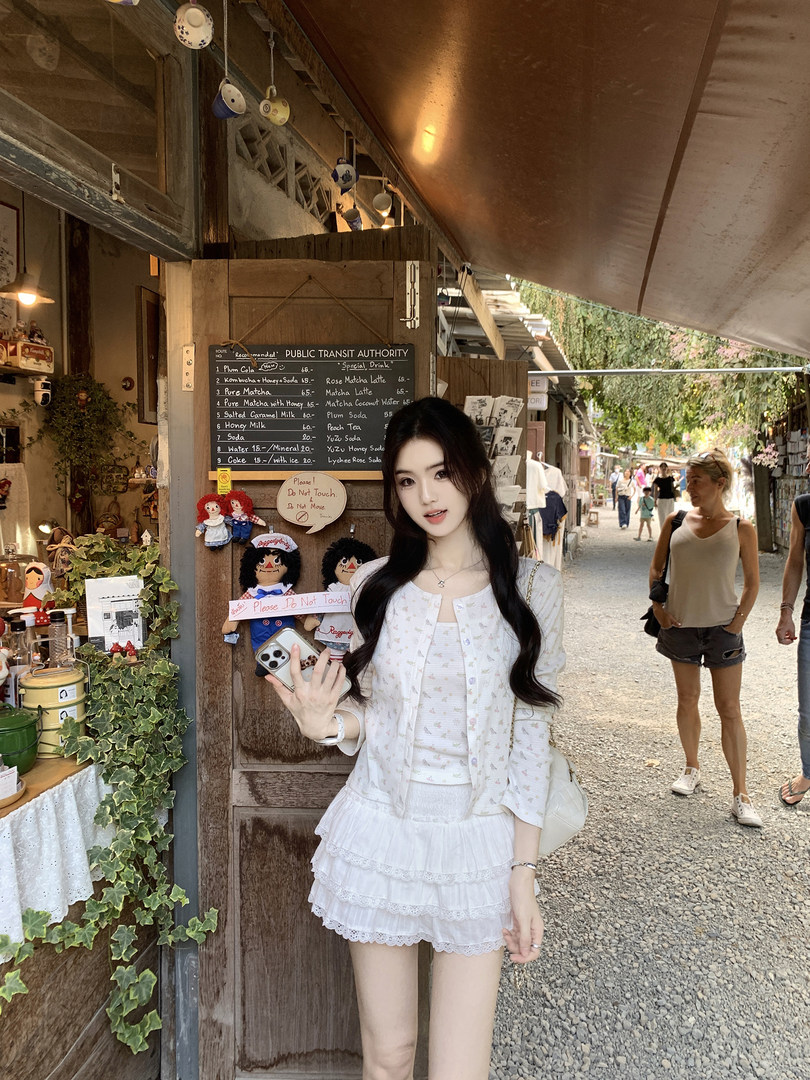 Woman wearing a white floral cardigan and ruffled shorts set, standing in front of a shop with a menu board.