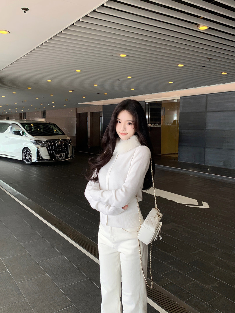 Woman wearing a white ribbed cardigan with faux fur collar, white pants, and carrying a white quilted chain bag, standing in a modern covered driveway with a white van in background.