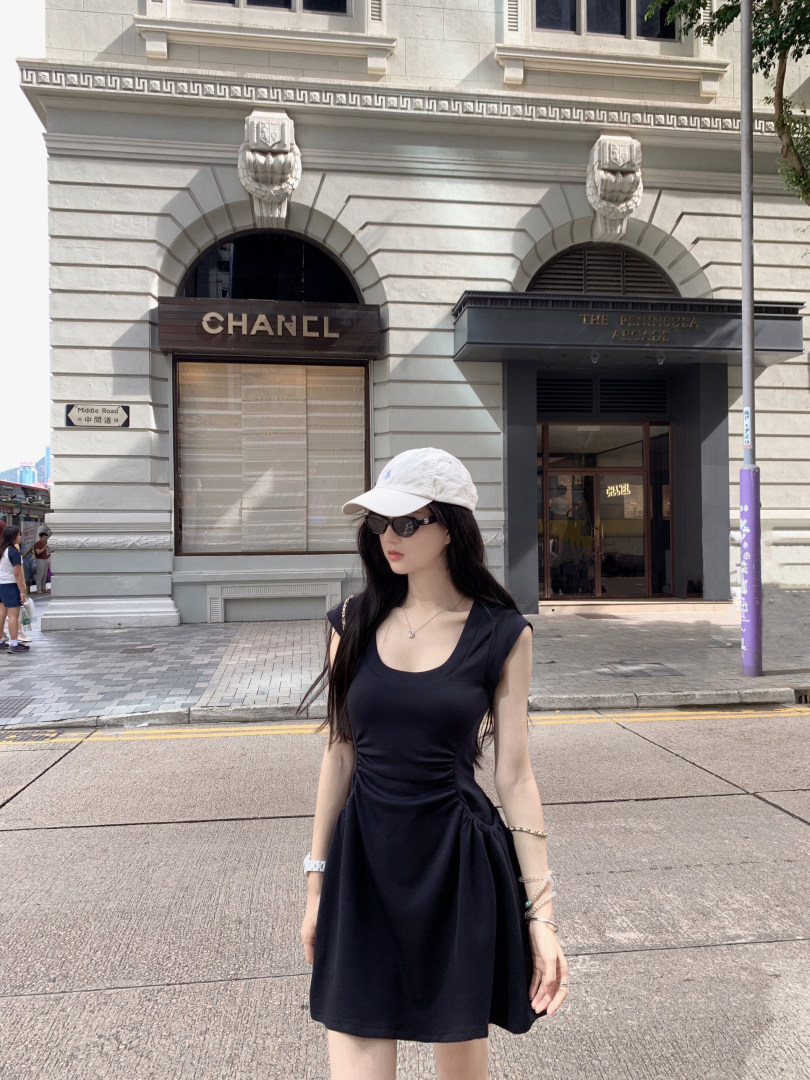 Woman wearing black sleeveless dress, white cap, and sunglasses standing on a city street
