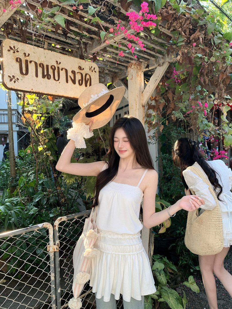 Woman wearing white lace-trimmed camisole top and holding straw hat under floral pergola