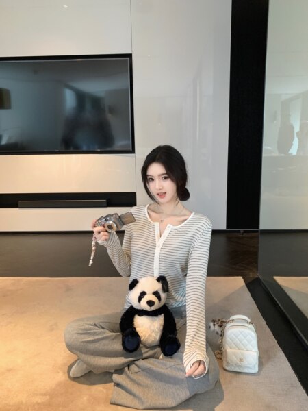 Woman sitting on floor wearing striped long-sleeve top and gray pants, holding camera with panda plush and white bag nearby