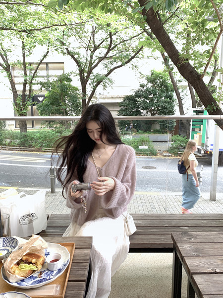 Woman wearing a light pink knit cardigan and white skirt sitting at an outdoor table with food and a camera