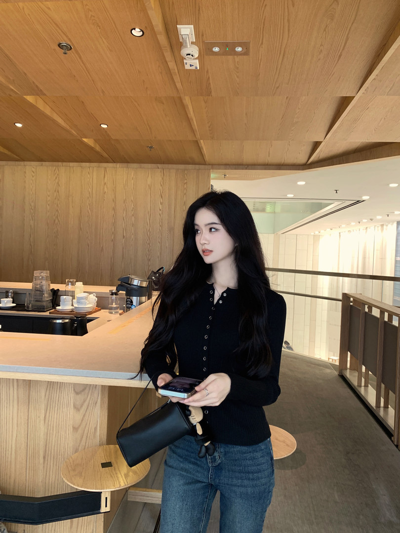 Woman wearing black ribbed cardigan and jeans, holding a phone and black bag in a modern wooden interior.