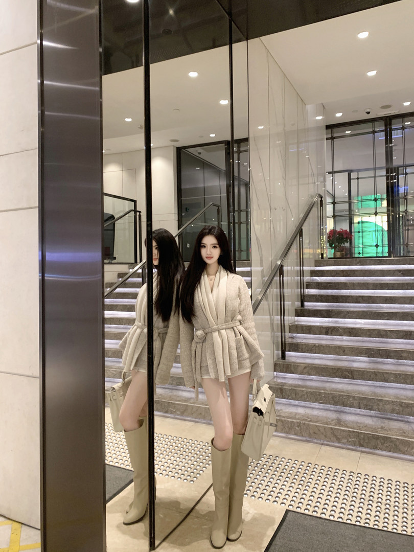Woman wearing beige belted coat and matching knee-high boots, standing in front of mirrored elevator with staircase background