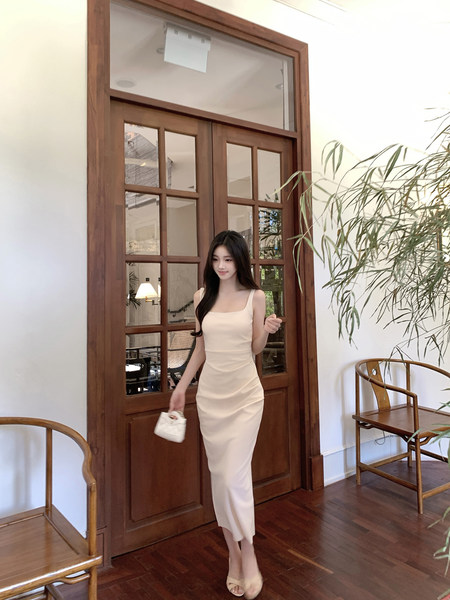 Woman wearing a beige sleeveless midi dress, holding a small white handbag, standing indoors near a wooden door.